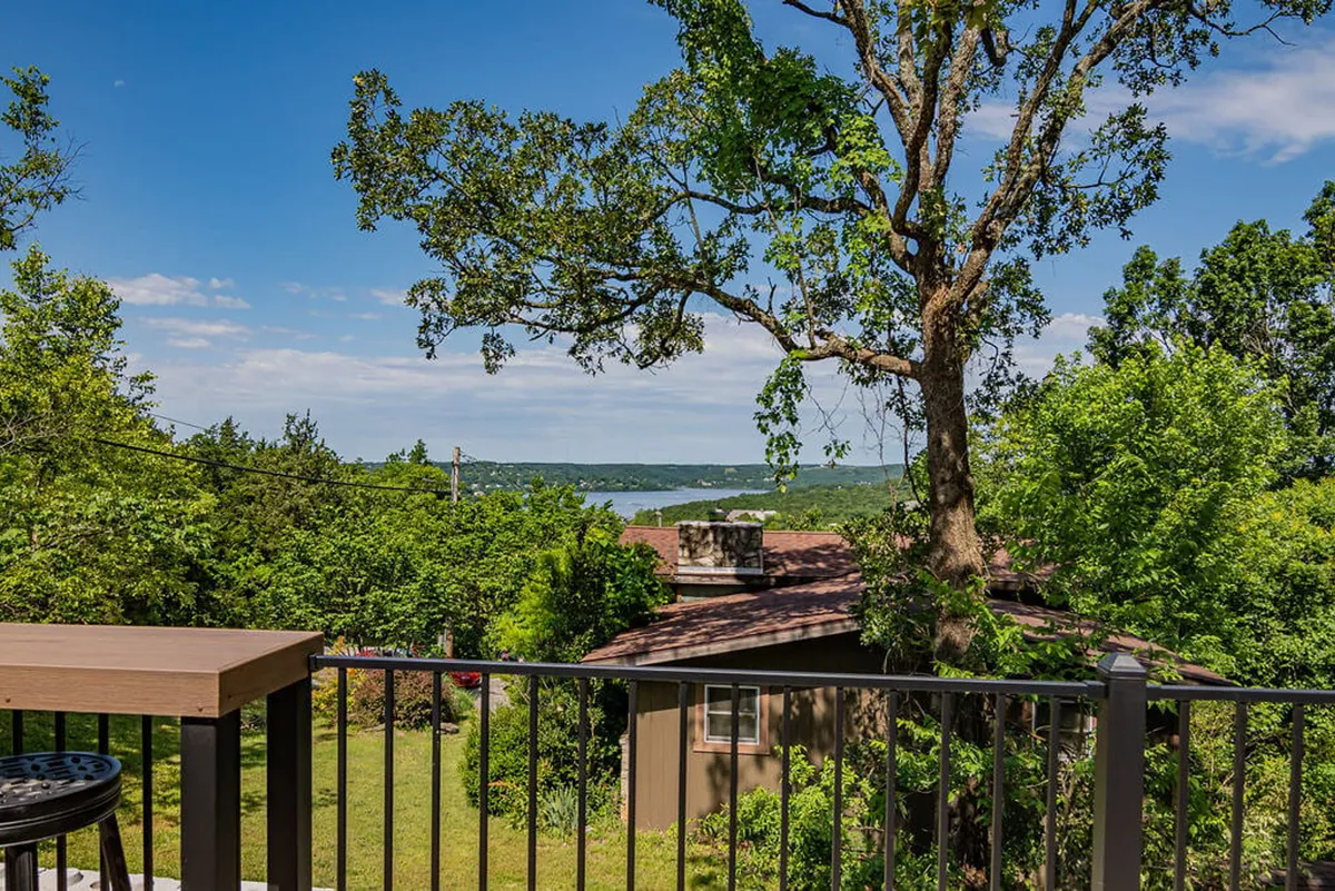 A sweeping view of Table Rock Lake framed by lush greenery and tree branches, with a deck area featuring comfortable seating. The scene invites relaxation outdoors, hosting gatherings, enjoying coffee, and bird watching, all set against a serene lakeside backdrop.