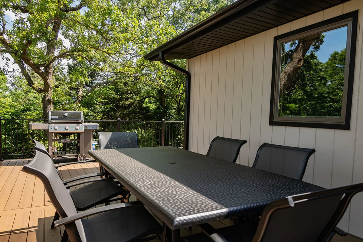 An exterior view of a deck adjacent to the kitchen, featuring a dining table, grill, and lush surrounding trees. The setup suggests effortless transitions from indoor cooking to al fresco dining, with direct access from the kitchen to the upper deck.