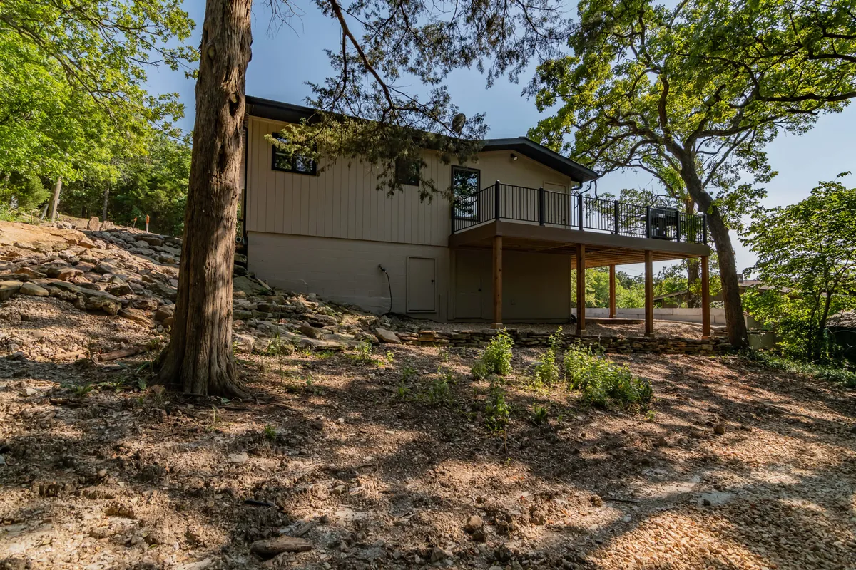 This exterior image shows the home nestled among mature trees, with a spacious deck area extending from the house. The setting highlights the natural tranquility of the Ozarks, with the deck positioned to catch the sunset and offer views of the surrounding landscape.