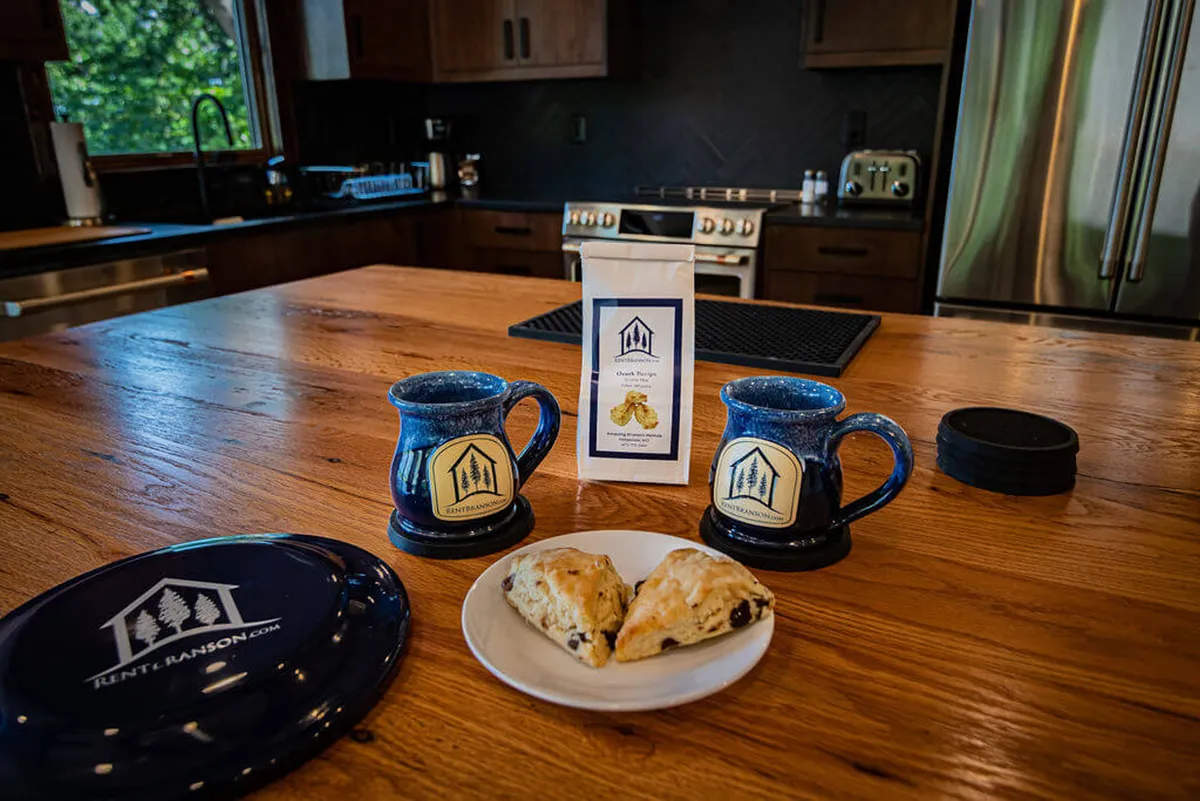 A close-up of a wooden kitchen island set for breakfast, with ceramic mugs, scones on a plate, and a coffee package. The setup evokes leisurely mornings and shared meals, highlighting the kitchen as a hub for cherished rituals.
