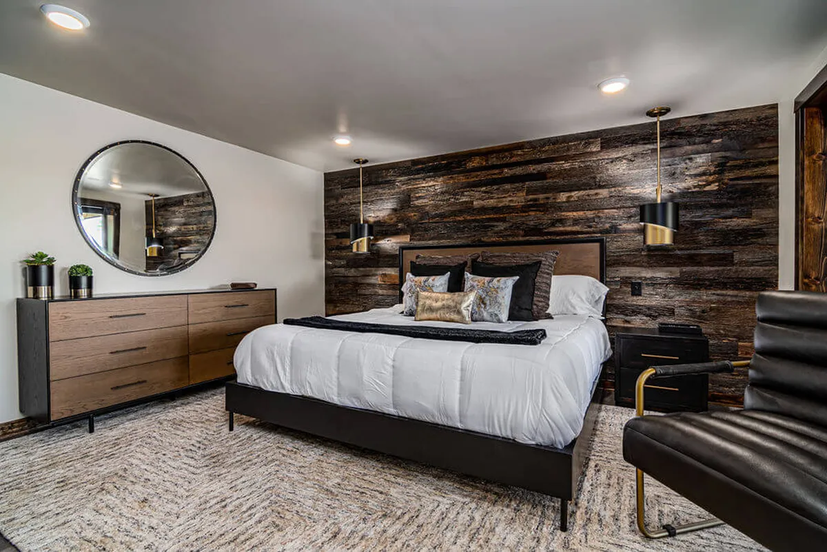A modern, inviting bedroom with a bed dressed in plush decorative pillows, a round mirror, warm wood accent wall, and a nightstand with a lamp. The room is softly lit and exudes a peaceful, restful ambiance, perfect for relaxation or a lazy morning.