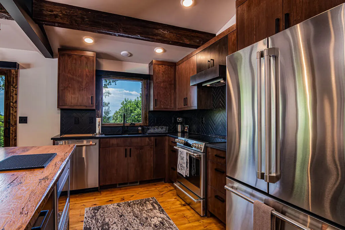 A bright, modern kitchen featuring stainless steel appliances, warm wooden cabinetry, a striking herringbone backsplash, and a large window flooding the space with natural light. The kitchen is set up for both cooking and entertaining, suggesting an inviting atmosphere ideal for family gatherings or casual breakfasts.