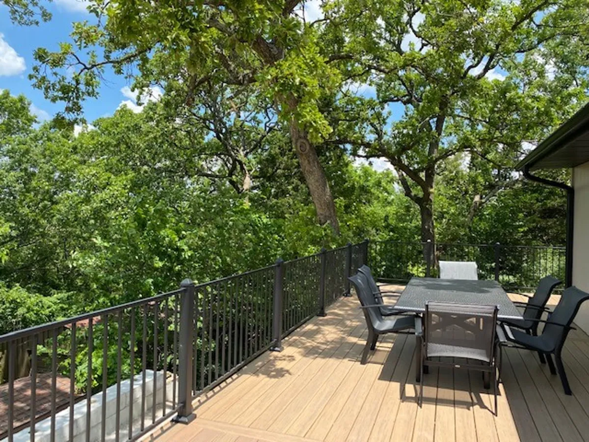 An outdoor dining area with a large table and chairs set on a wooden deck, surrounded by greenery and bordered by a railing. The scene is set for sunset dinners, stargazing, or simply gathering with friends and family in a peaceful, natural environment.