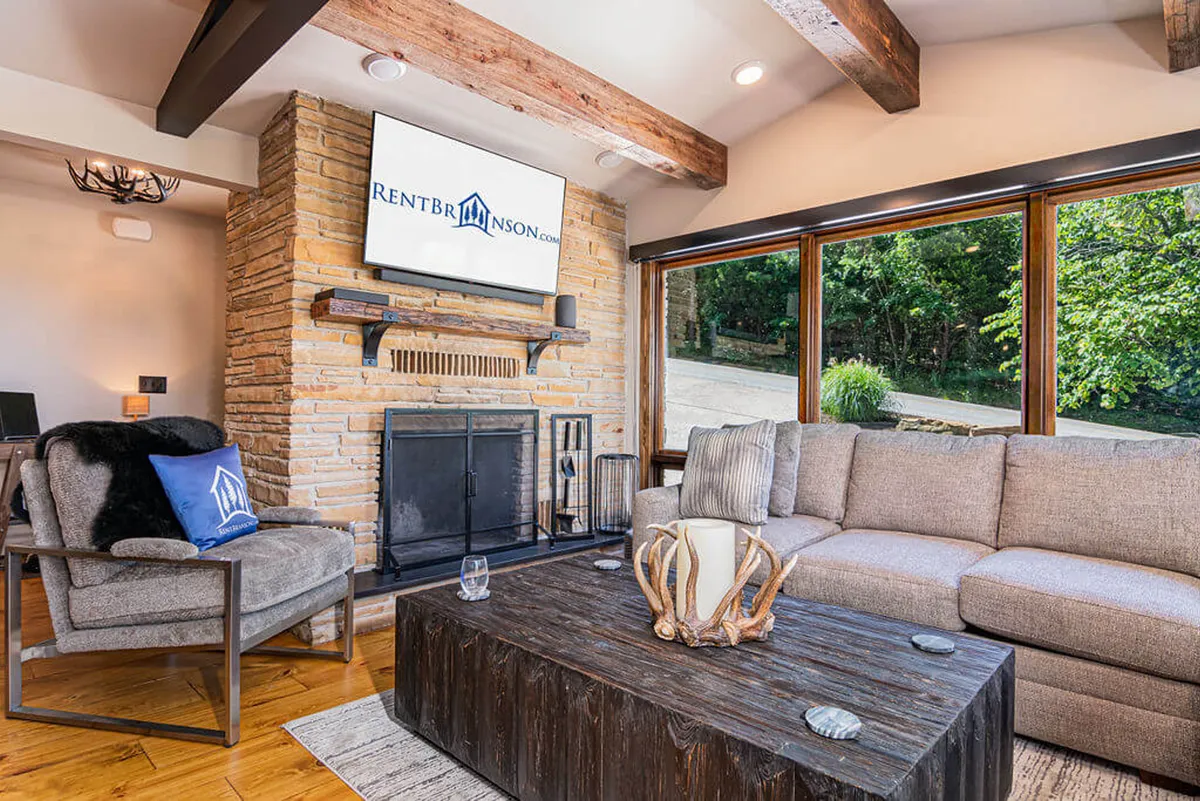 A welcoming great room with a plush sectional sofa facing a stone fireplace, large windows that frame scenic outdoor views, a central coffee table, and a wall-mounted high-definition smart TV. Designer furnishings and a mix of cozy textures create an inviting social atmosphere.
