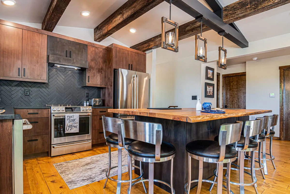 A view of the kitchen centered on a custom island with bar stools, surrounded by stainless steel appliances and wooden beams overhead. The setting shows the seamless flow between cooking, dining, and casual socializing, ideal for both meal prep and shared moments.