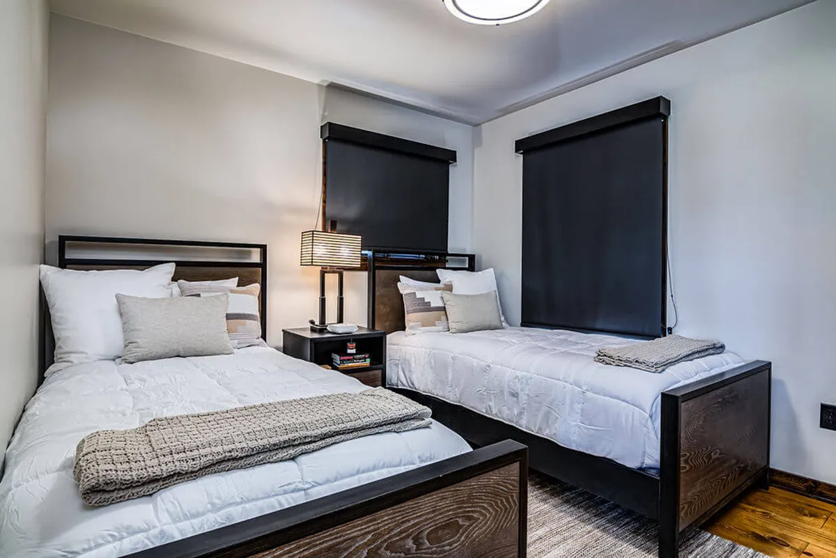 A serene twin bedroom with two twin beds, a nightstand with a lamp, and decorative pillows. Light filters through window blinds, creating a restful setting ideal for families or friends seeking comfort and privacy.