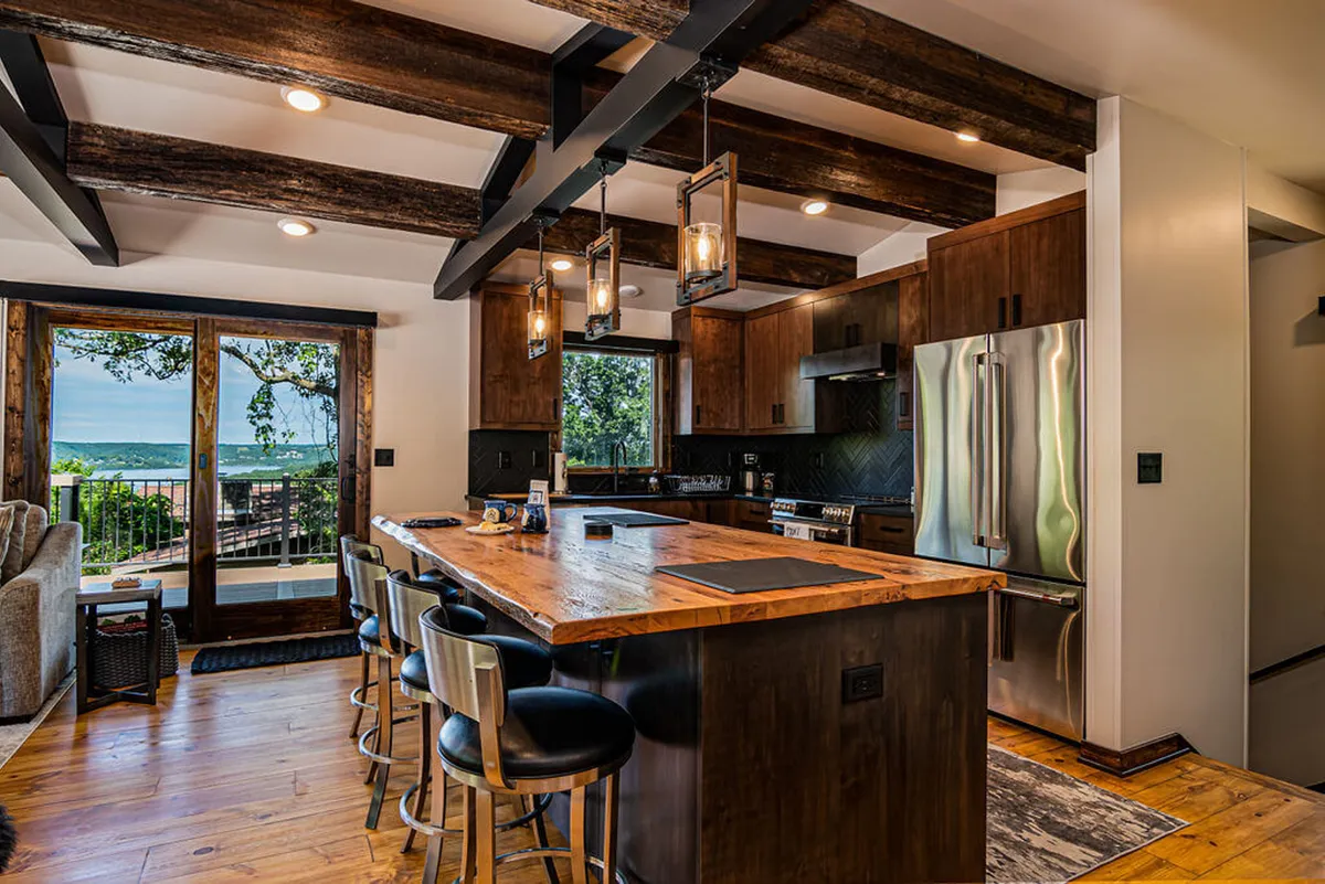 The image showcases a warm, inviting kitchen with a large island perfect for family gatherings, modern high-end appliances, striking wooden beams overhead, and expansive windows offering panoramic views of Table Rock Lake. Sunlight pours in, highlighting the blend of mid-century modern architecture and natural materials, with the outside scenery taking center stage through the glass.