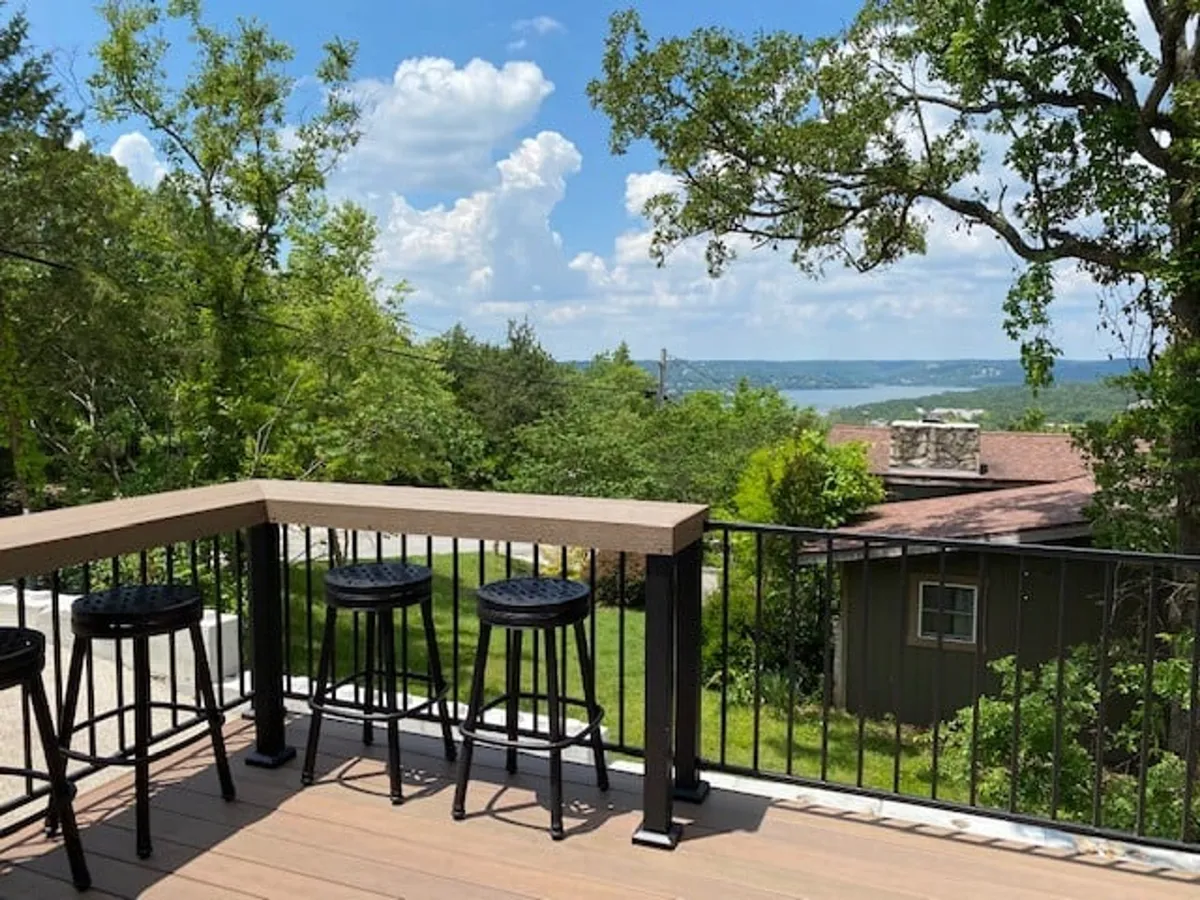 A spacious deck with bar stools and a deck railing offers expansive views of the Ozark landscape. People are depicted enjoying refreshments, socializing, and relaxing outdoors, capturing the essence of morning coffees, casual conversations, and evening gatherings.