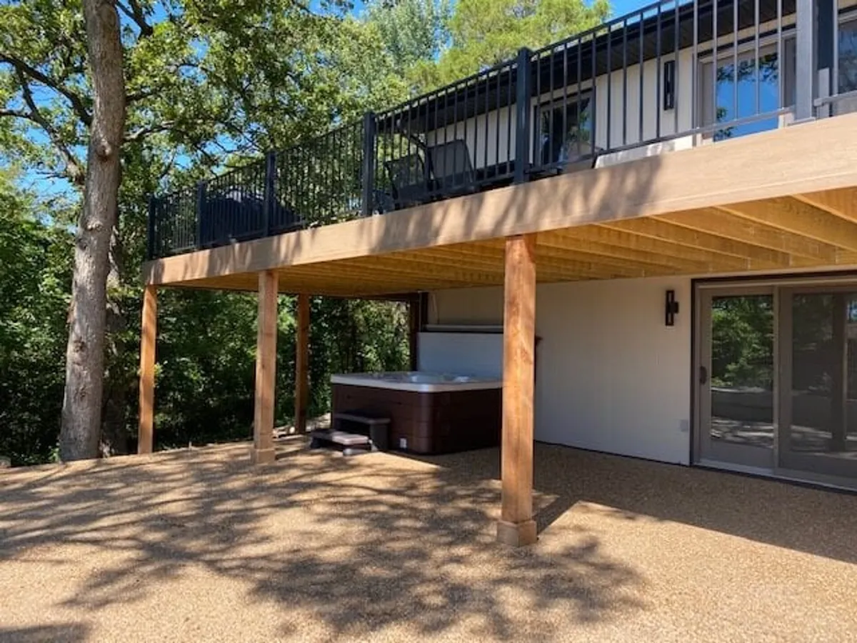 The exterior showcases a spacious deck with a hot tub, wooden beams, and balcony chairs, set against a backdrop of trees. The space is perfect for relaxing in the hot tub, enjoying outdoor seating, and hosting gatherings, all accompanied by tranquil surroundings.